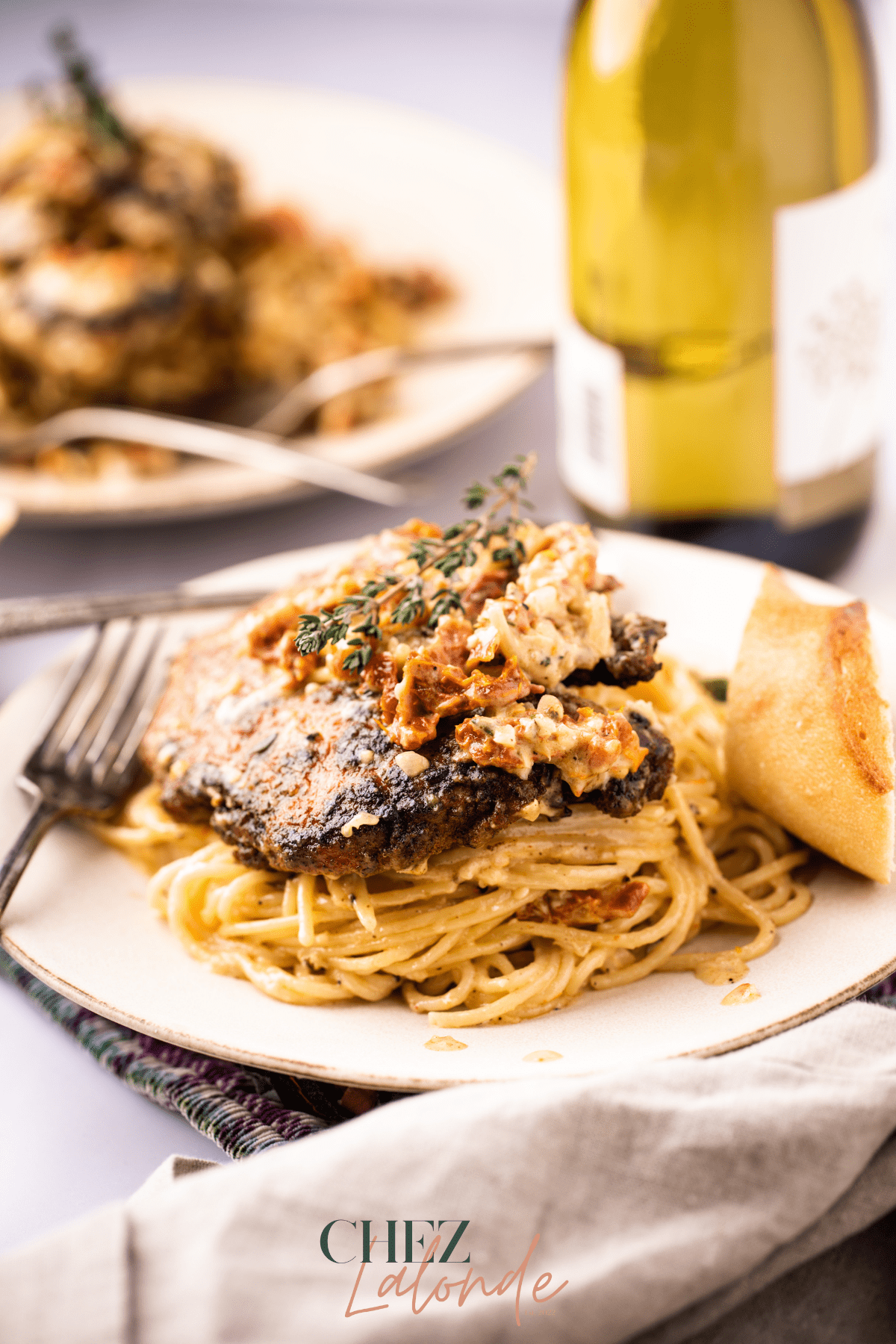 Sun-Dried tomato pasta with pan-seared chicken breast on a white plate. There a a bottle of white wine, vintage silverware, and bread