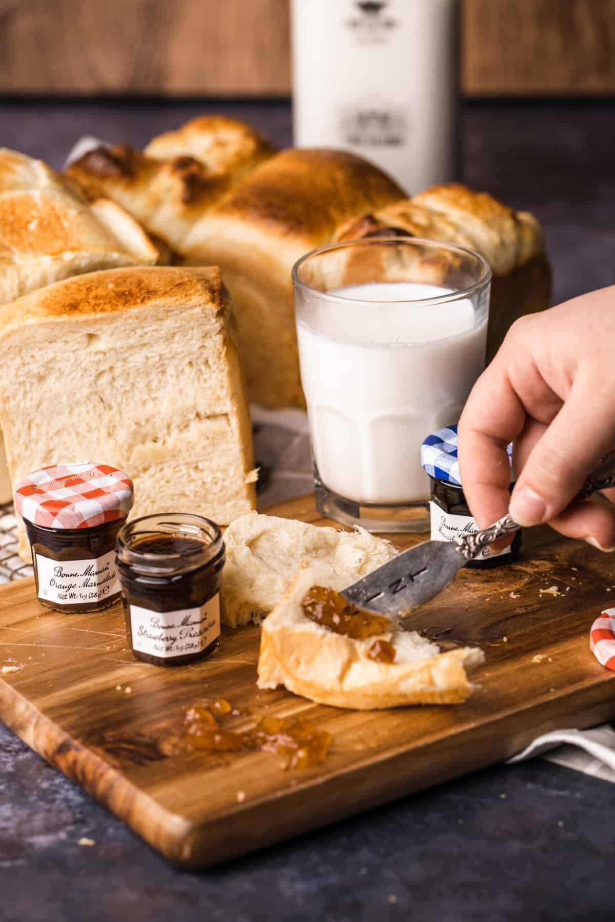a girl is spreading jam onto a piece of Japanese milk bread with 2 other loaf of bread in the back ground. There are also a glass of milk and 3 small jars of jam. 