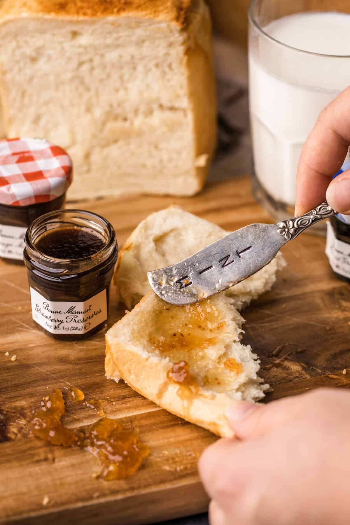 a girl is spreading jam onto a piece of Japanese milk bread with 2 other loaf of bread in the back ground. There are also a glass of milk and 3 small jars of jam.
