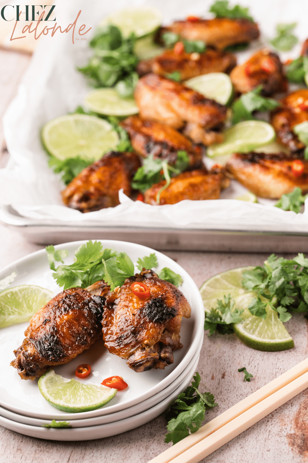Chez Lalonde Lemongrass Chicken Wings sitting on a cooking tray and 2 wings are on a stacks of small white plates. There are some limes, cilantro and chopsticks