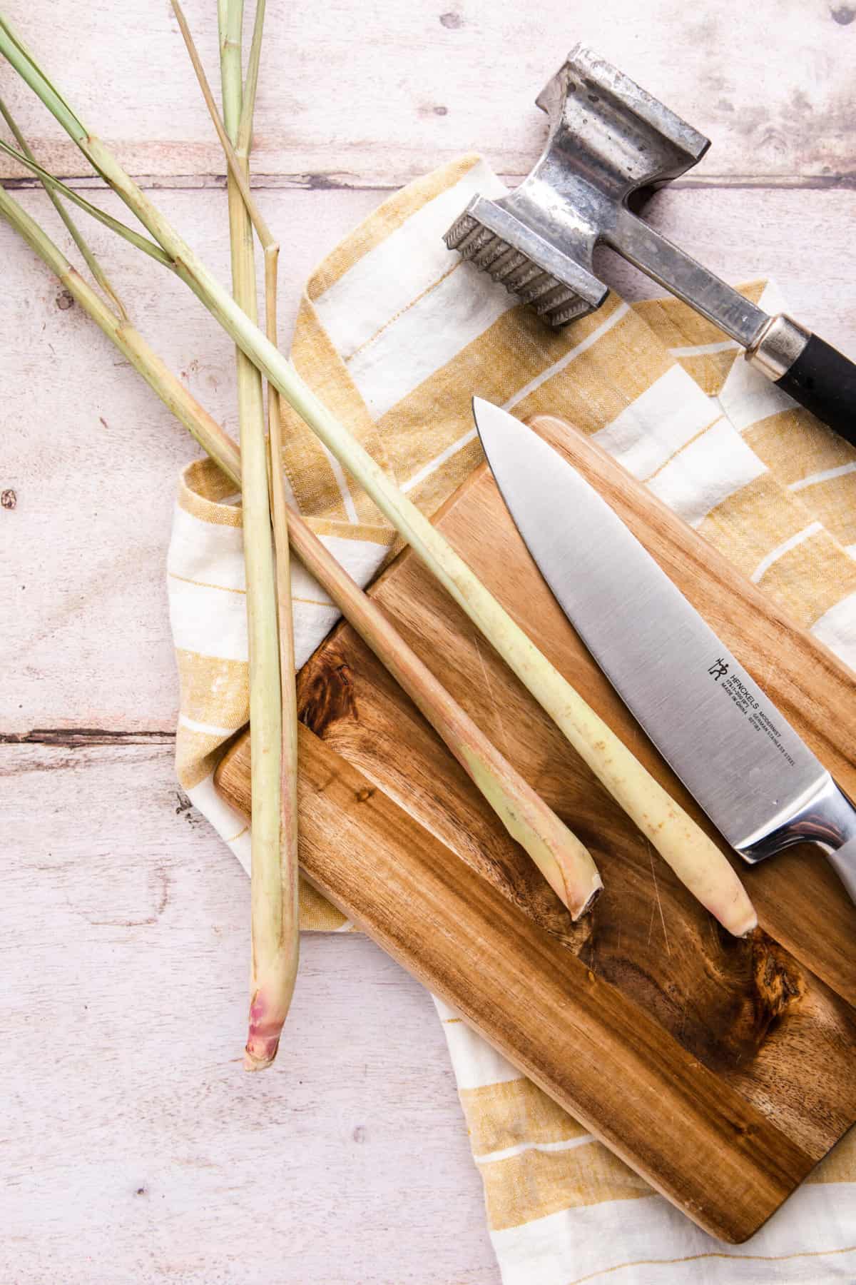 Chez Lalonde 3 lemongrass sitting on top of a wooden chopping board. Knife on the right hand side with a meat tenderizer on top right corner