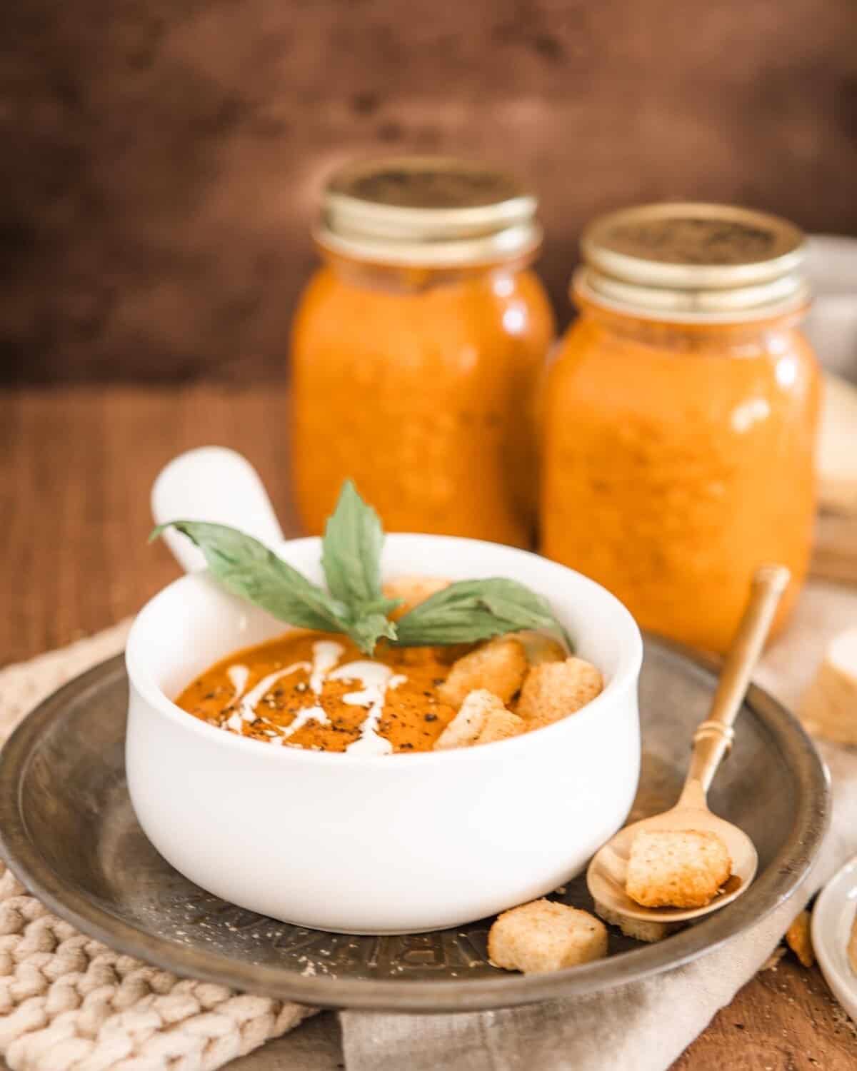 Roasted tomato soup with croutons, basil, and cream. Soup is contained in a white bowl with short handle served on a silver plate with a golden spoon. There are 2 jars of soup in the background
