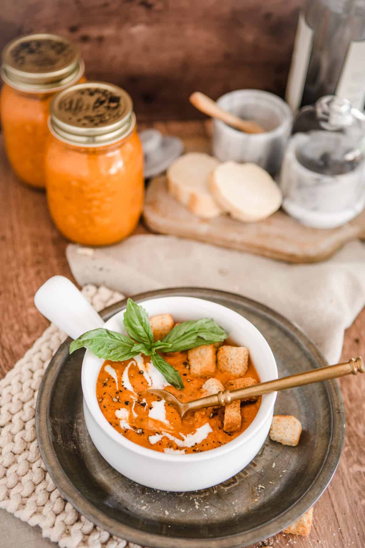 Creamy Roasted Tomato and Basil Soup in a white bowl with golden spoon on a vintage plate.  Also have 2 jars of canned soup, bread. 
