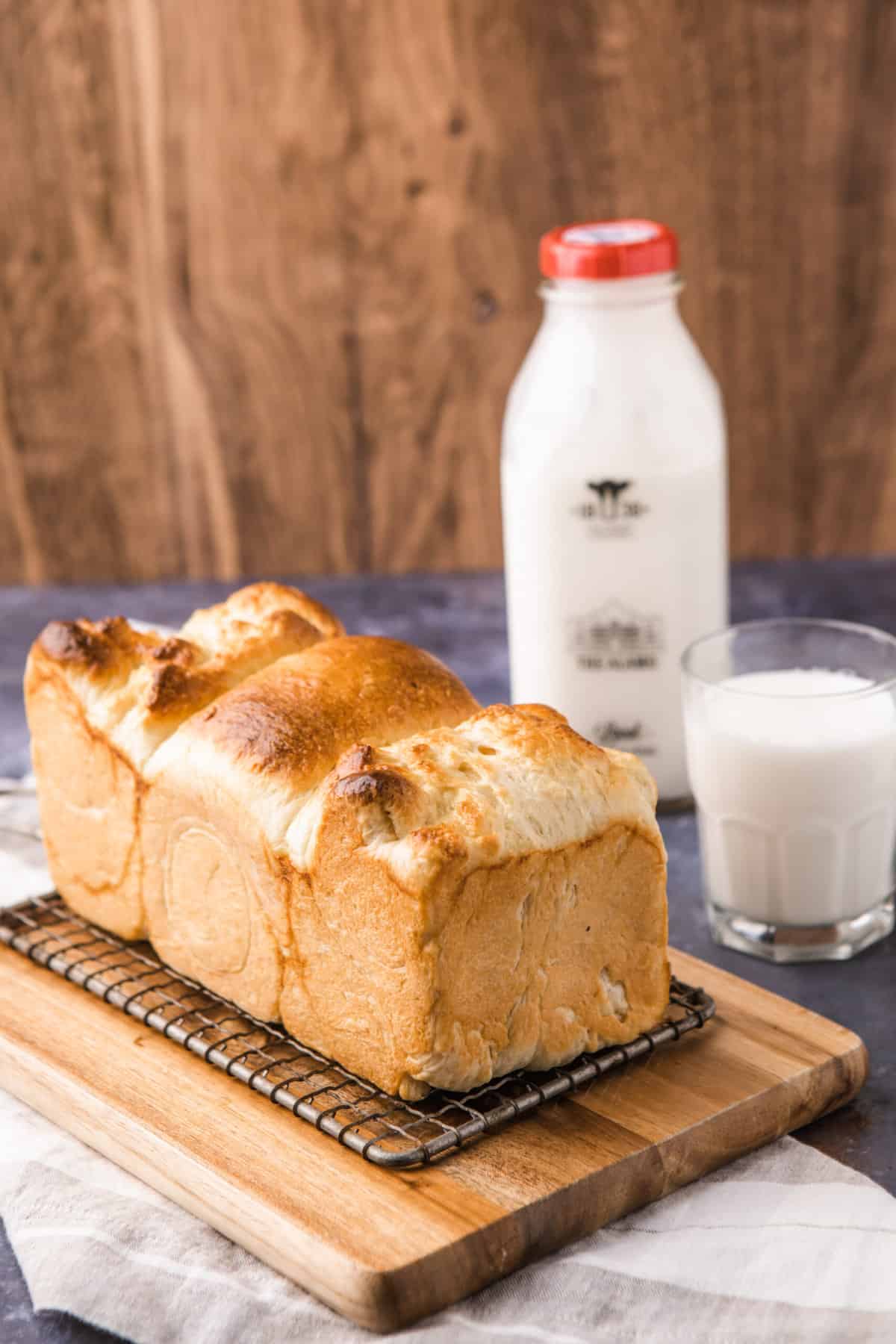 a loaf of japanese milk bread sitting on a small cutting board. a bottle of milk and a glass of milk are in the background. 