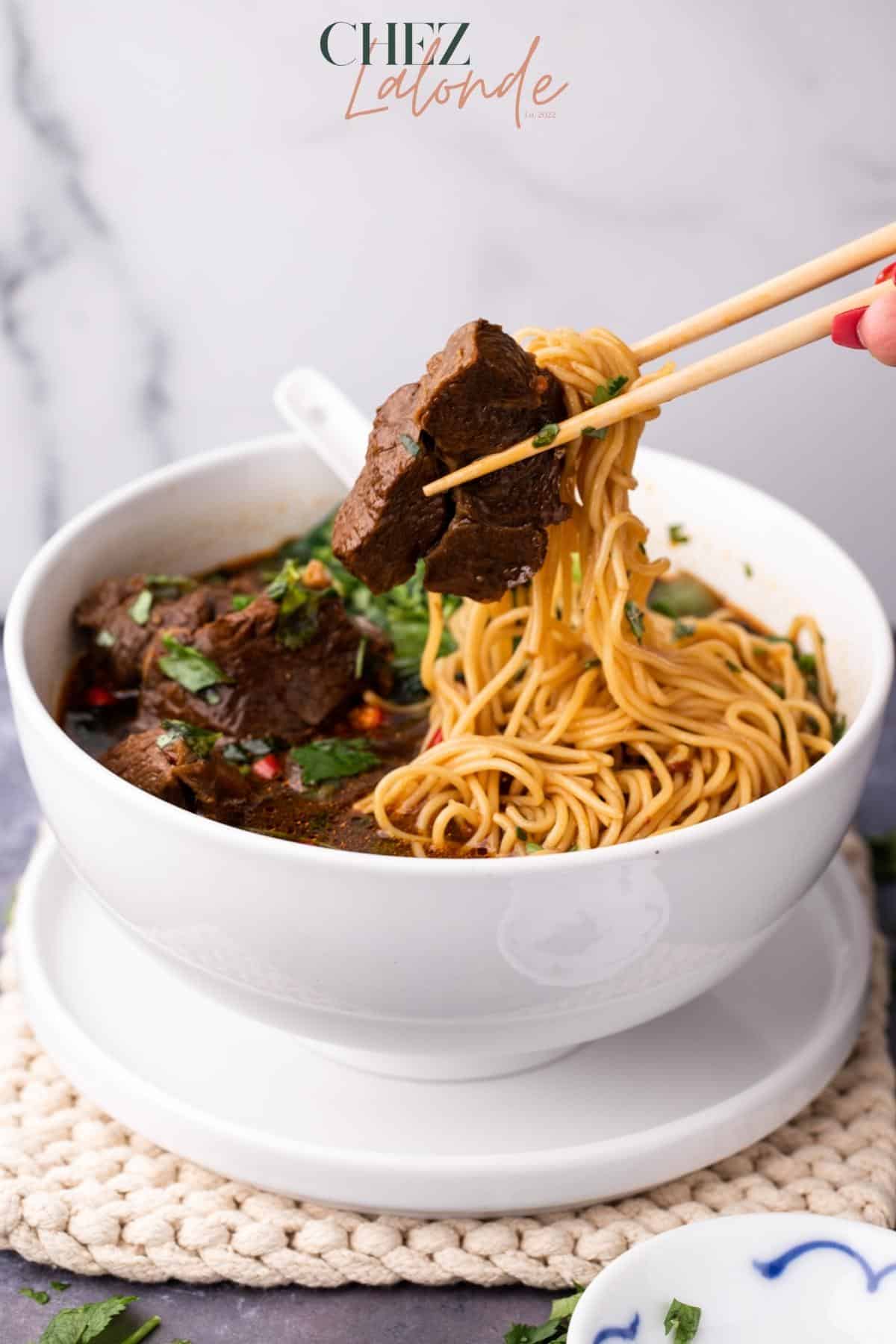 Taiwanese spicy beef noodle soup in a white bowl. A pair of chopsticks is picking up a piece of tender beef shank and some wheat noodles. The bowl seats on top of a round saucer.