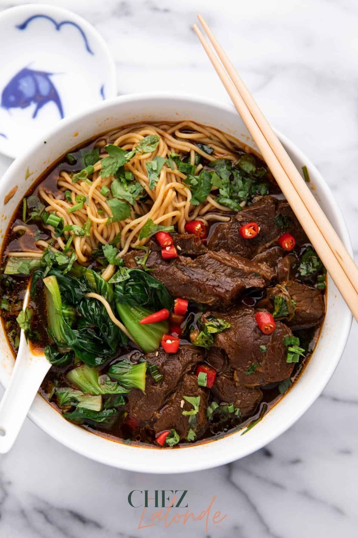 Delicious Taiwanese spicy beef noodle soup contained in a white bowl. It is with a small sauce dish, chopsticks and Chinese spoon