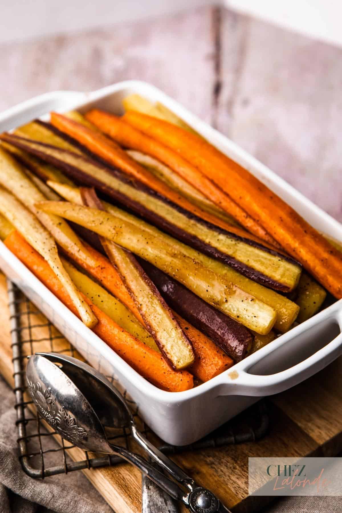 Maple Roasted rainbow carrots in a white baking tray.  The tray is sitting on top of the cutting board and there is a thong sitting on the side. 
