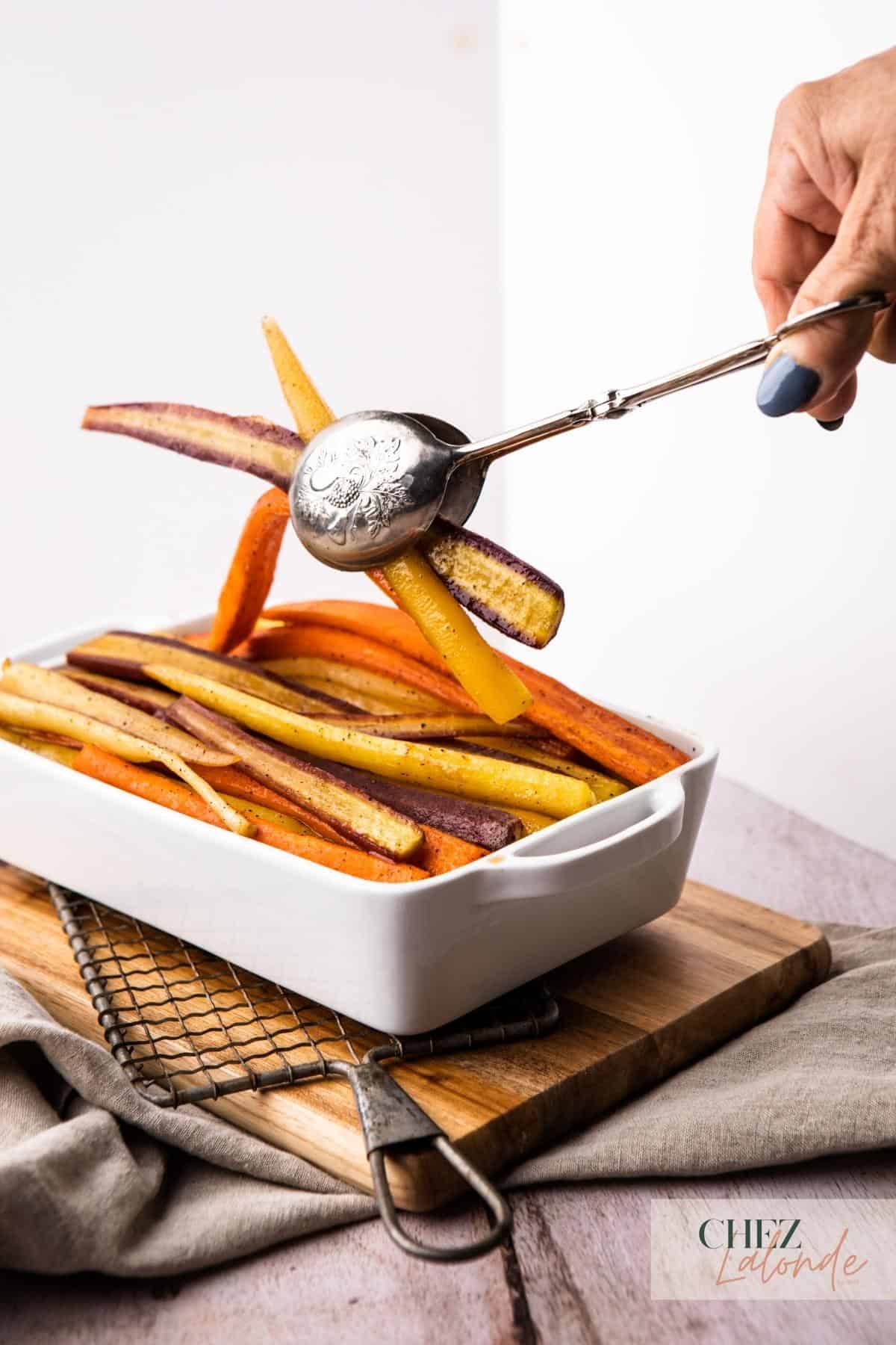 Maple Roasted rainbow carrots in a white baking dish.  A woman's hand is using a vintage thong to grab a few pieces up to serve for her guest.  