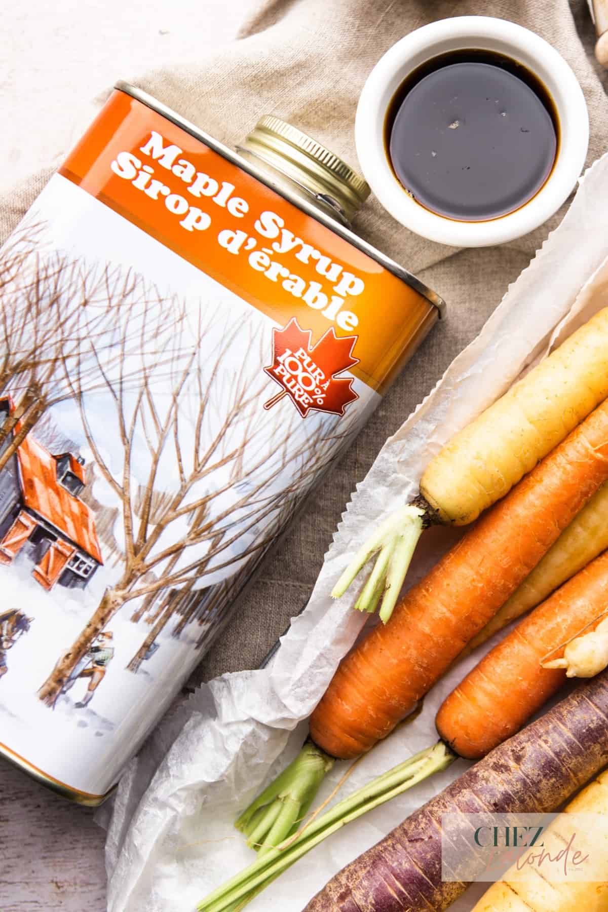 A can of real Canadian Maple Syrup and some pour out on a ramekin.  Some rainbow carrots are on the bottom corner of the picture.  