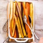 Maple Roasted Rainbow Carrots in a white baking dish. A hand is holding a thongs and grabbing some carrots. The baking tray is sitting on a cutting board.