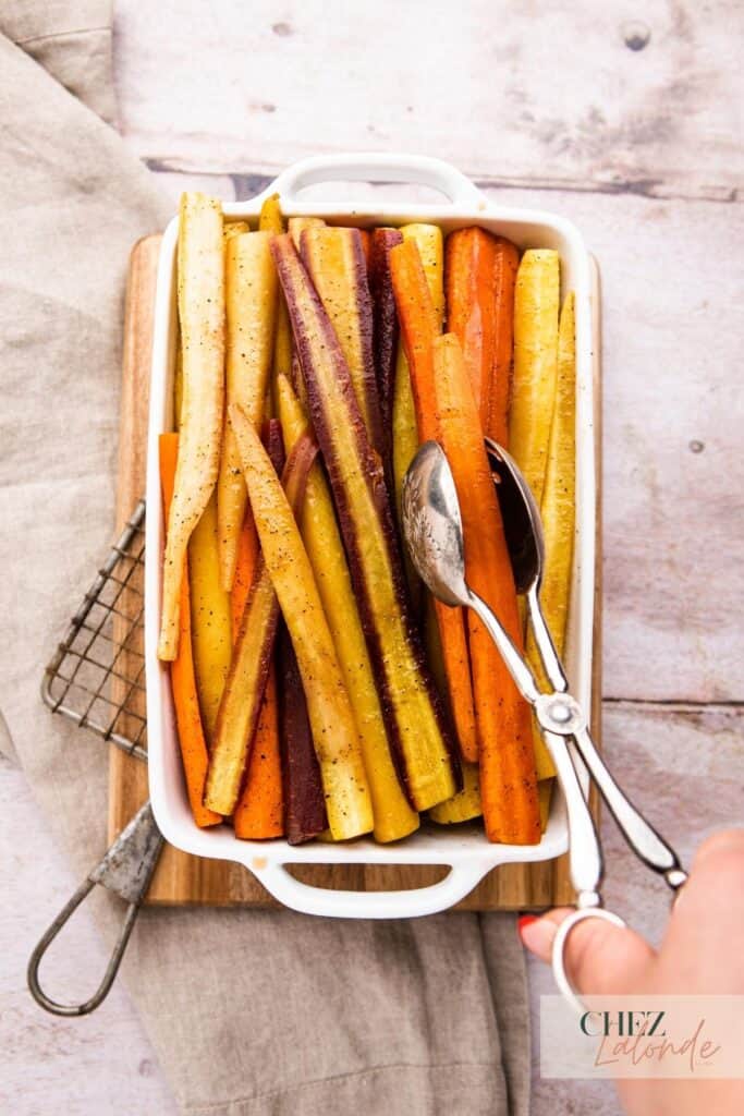 Maple Roasted Rainbow Carrots in a white baking dish. A hand is holding a thongs and grabbing some carrots. The baking tray is sitting on a cutting board.
