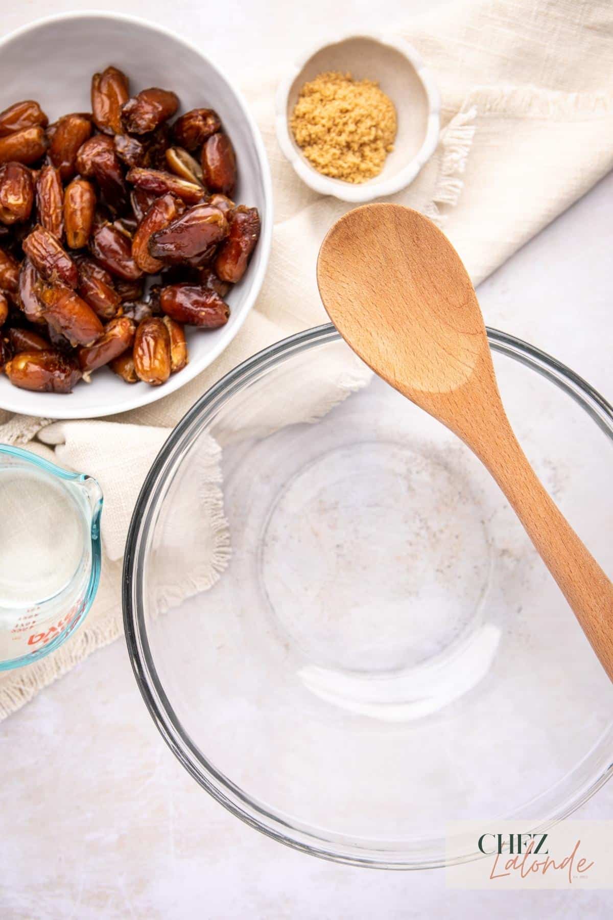 A bowl of dates, a glass mixing bowl, and a wooden spoon on a table.