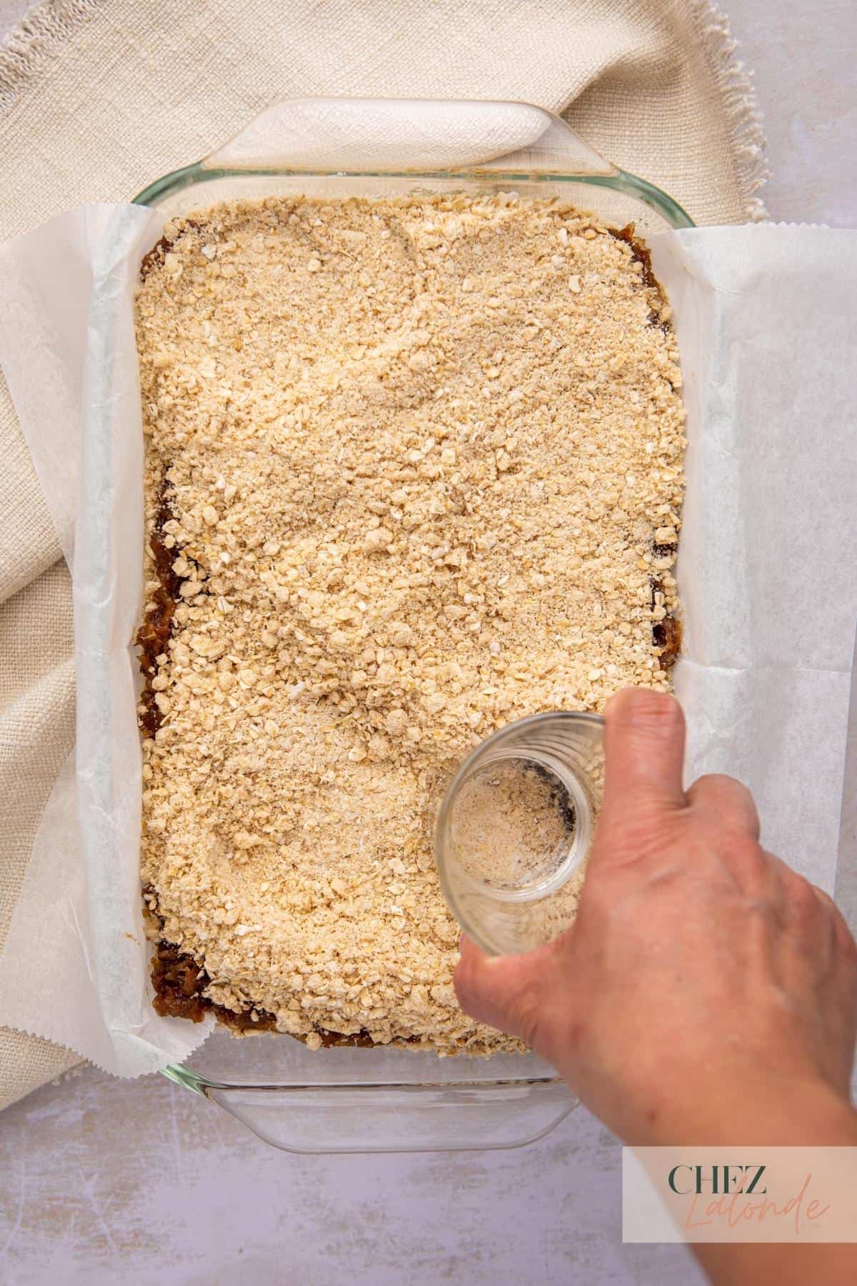 A baking dish with layers of date filling and oatmeal topping. A hand is using a glass bottom to press down the toppings.