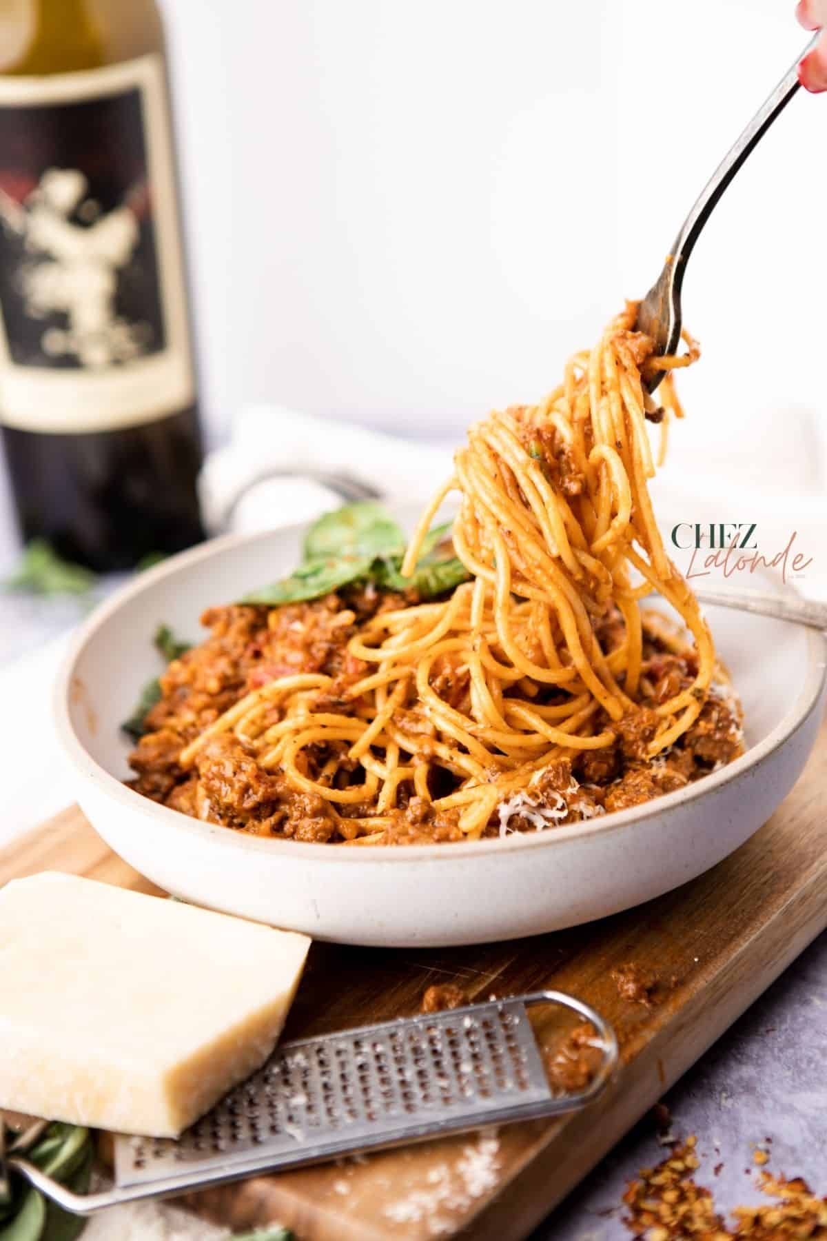 a plate of spaghetti with meat sauce sitting on a round bowl.  A hand with a fork is picking up some spaghetti.  A bottle of red wine is in the background.  Both white bowl and a wedge of cheese are sitting on top of a small cutting board. 