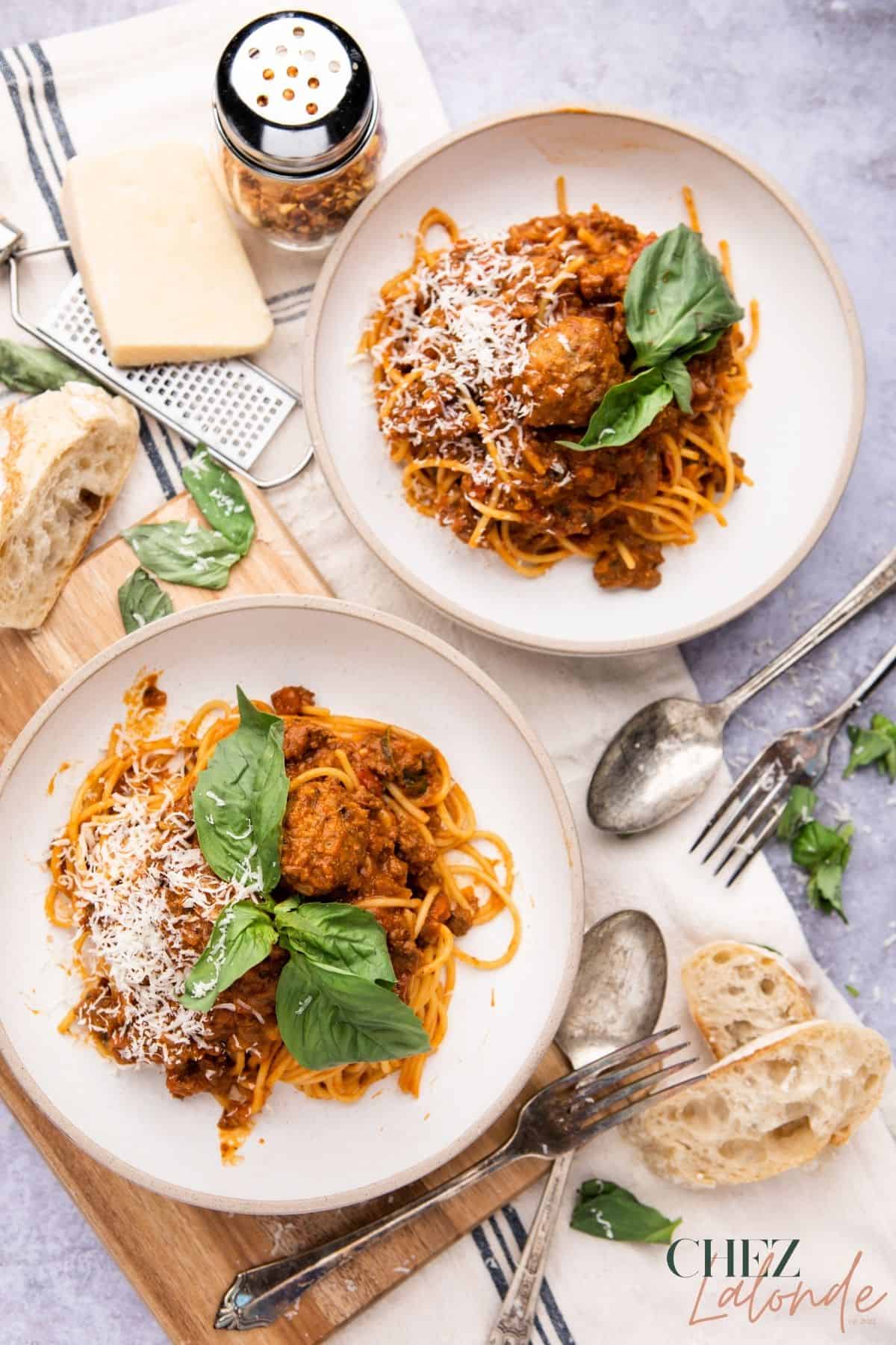 Two plates of spaghetti with meat sauce on  round white plates.  They are garnished with grated  with parmigiano-Reggiano cheese and fresh basil. There are slices of french baguette, a big wedge of cheese on the top left corner with a jar of red chili flakes. 