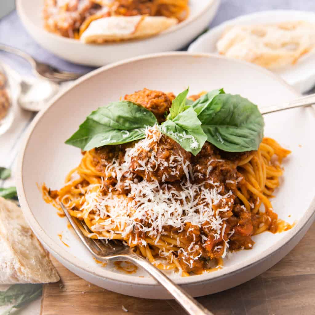 A plate of spaghetti with meat sauce that garnish with cheese and fresh basil. A fork and spoon are on the round white plate with the pasta.