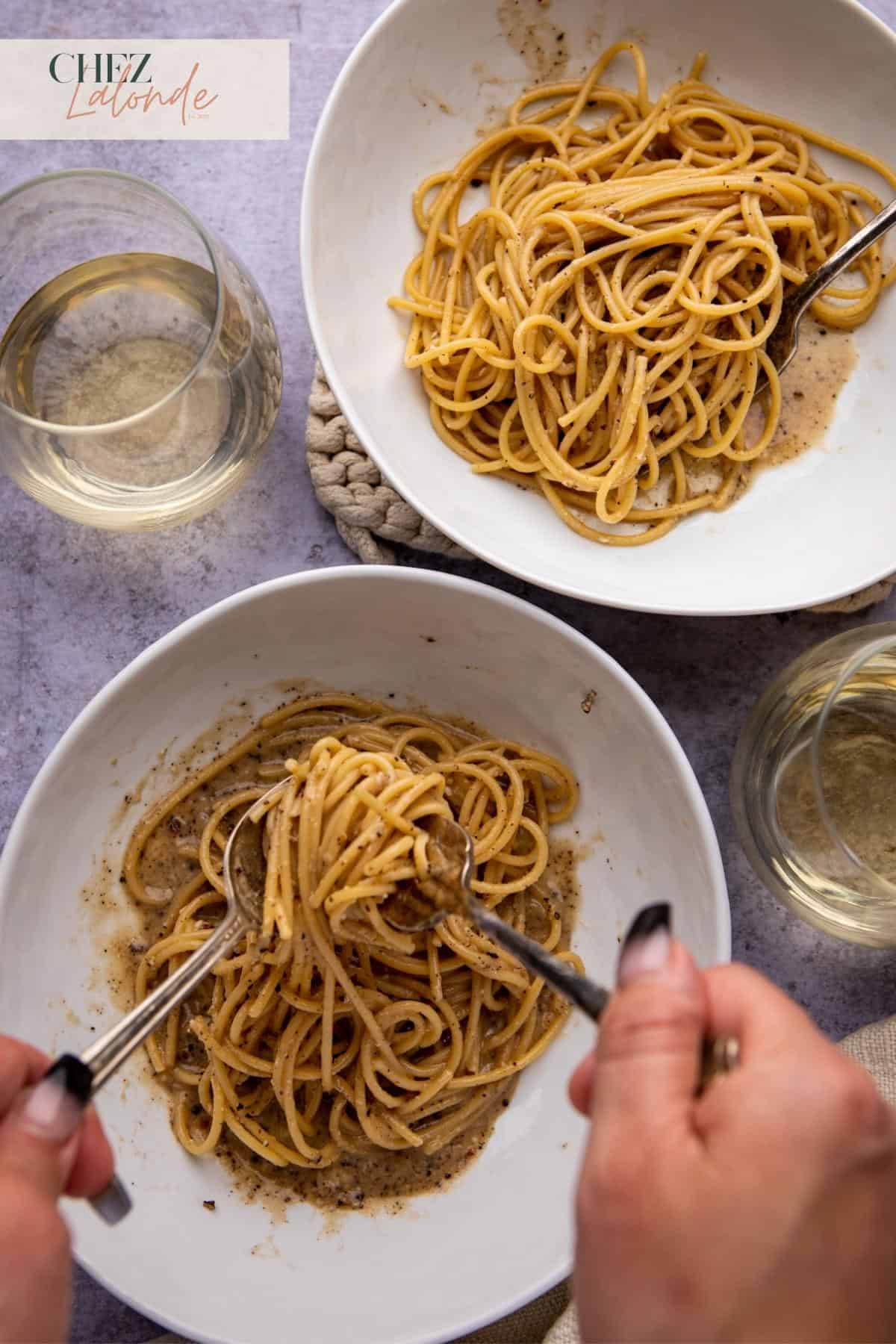 a woman is eating her cacio e pepe spaghetti with another plate on top right corner as well as 2 glasses of white wine. 