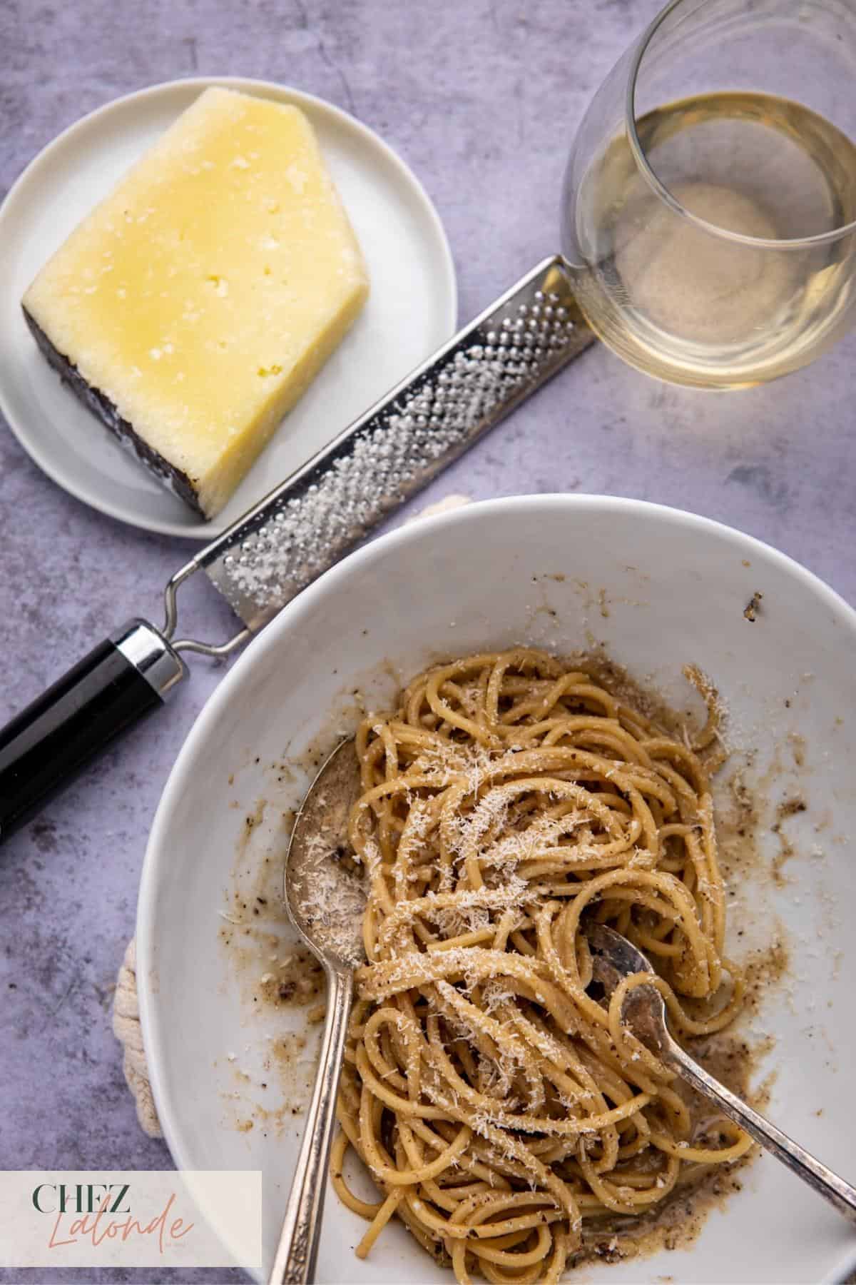A plate of Cacio e pepe spaghetti on a round white bowl with a microplane tool, a block of pecorino romano cheese and a glass of white wine. 