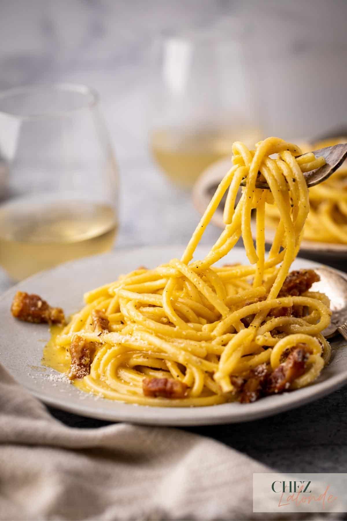 A fork is holding up some Italian Carbonara and there are another plate of Carbonara in the background with 2 glasses of white wine. 