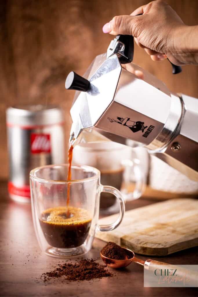 a hand is holding a moka pot and pouring fresh brewed coffee in a glass mug.