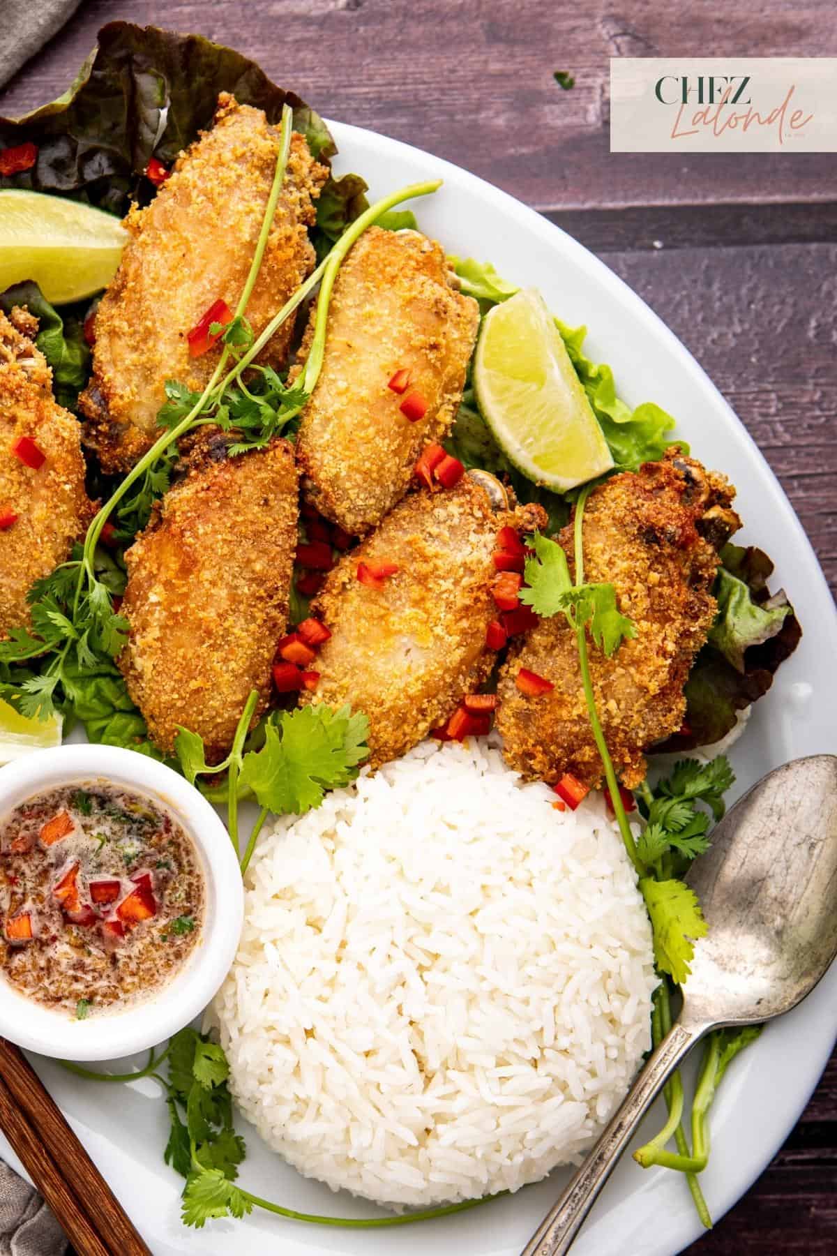 A plate of air fried chicken wings with white rice, vietnamese dipping sauce and garnished with cilantro, lime, and pepper. 