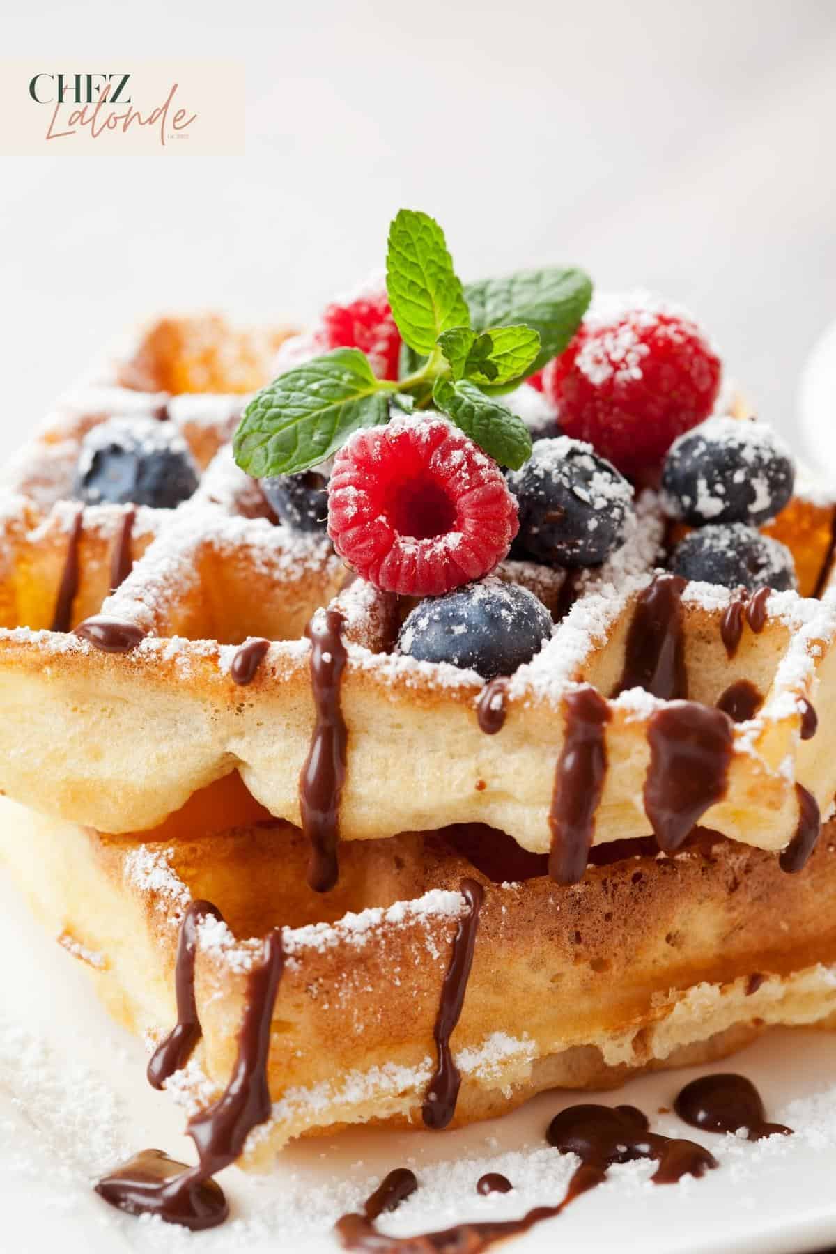 A stack of American Waffles with chocolate drizzle, berries and sugar powder. 