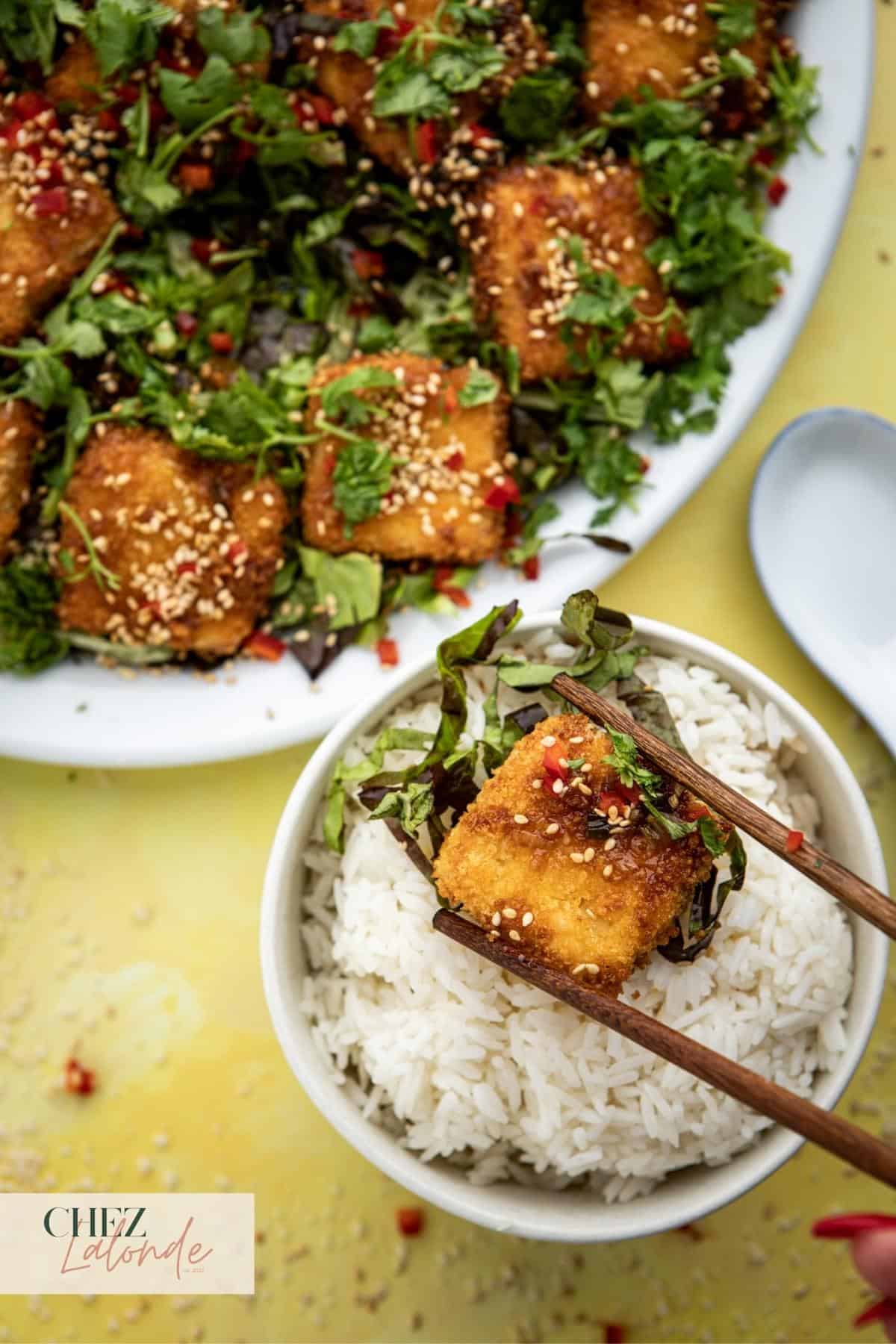 A pair of chopsticks are picking up a piece of air fried tofu and place it on top of a bowl of white rice. 