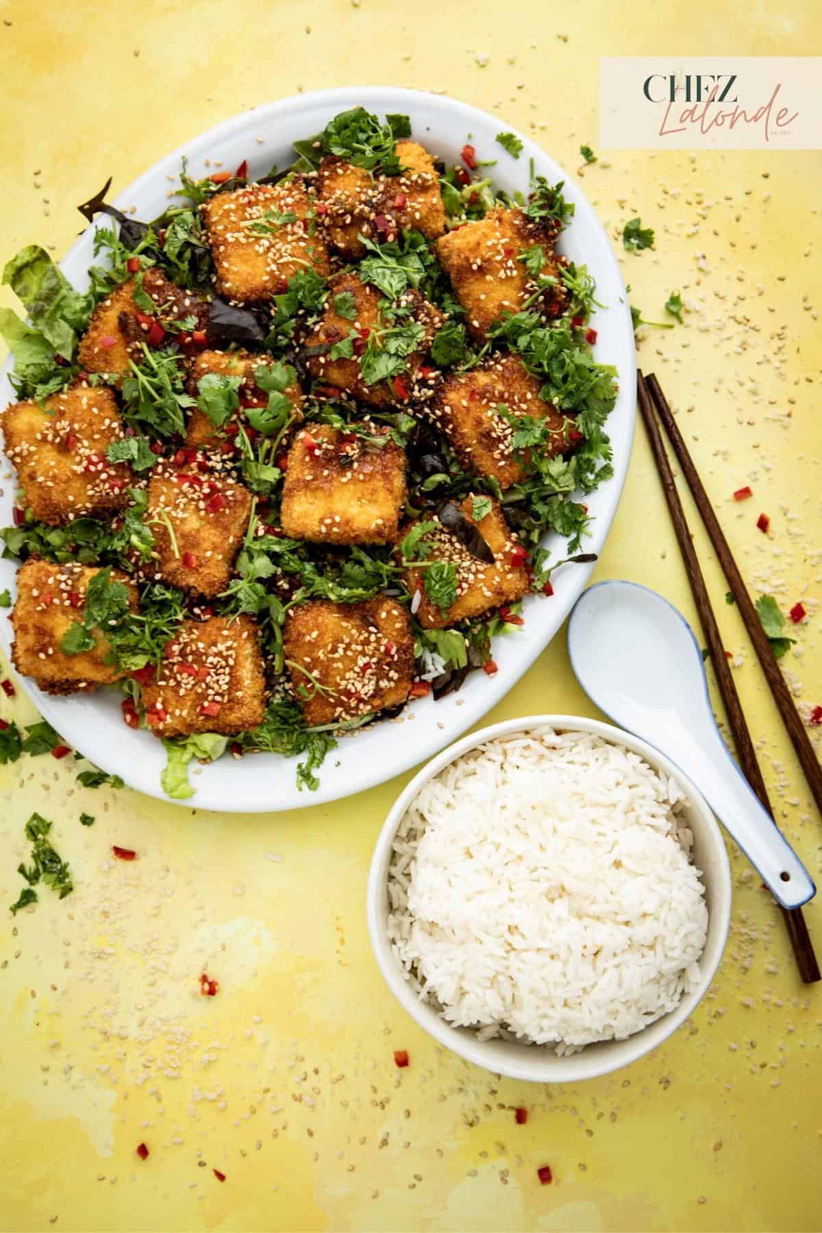 A plate of air fried tofu on a bed of shredding lettuce and white plate.  A bowl of steamed rice, spoon, and a pair of chopsticks are next to the plate of air fried tofu. 