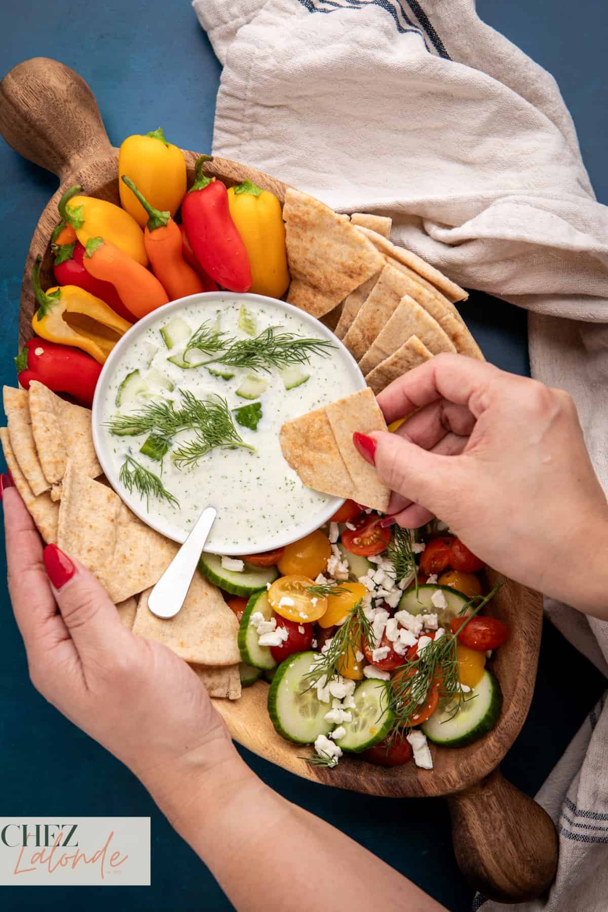 A woman is dipping some warm pita bread in a bowl of Tzatziki sauce. The wooden bowl contains Tzatiziki, fresh Greek Salad, warm pita bread and sweet peppers.