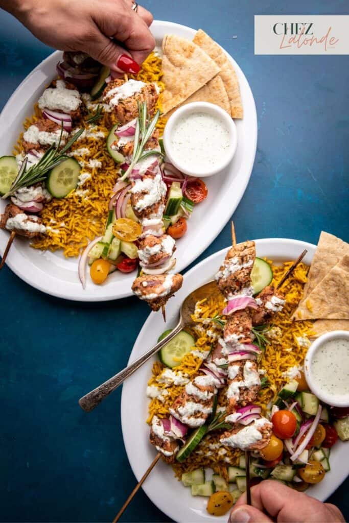 Here are 2 plates of Sous Vide Pork tenderloin Souvlaki served with tangy Tzatziki, Greek salad, Mediterranean rice and pita. 2 people are having this dish for dinner and their hands are picking up one of the skewer.
