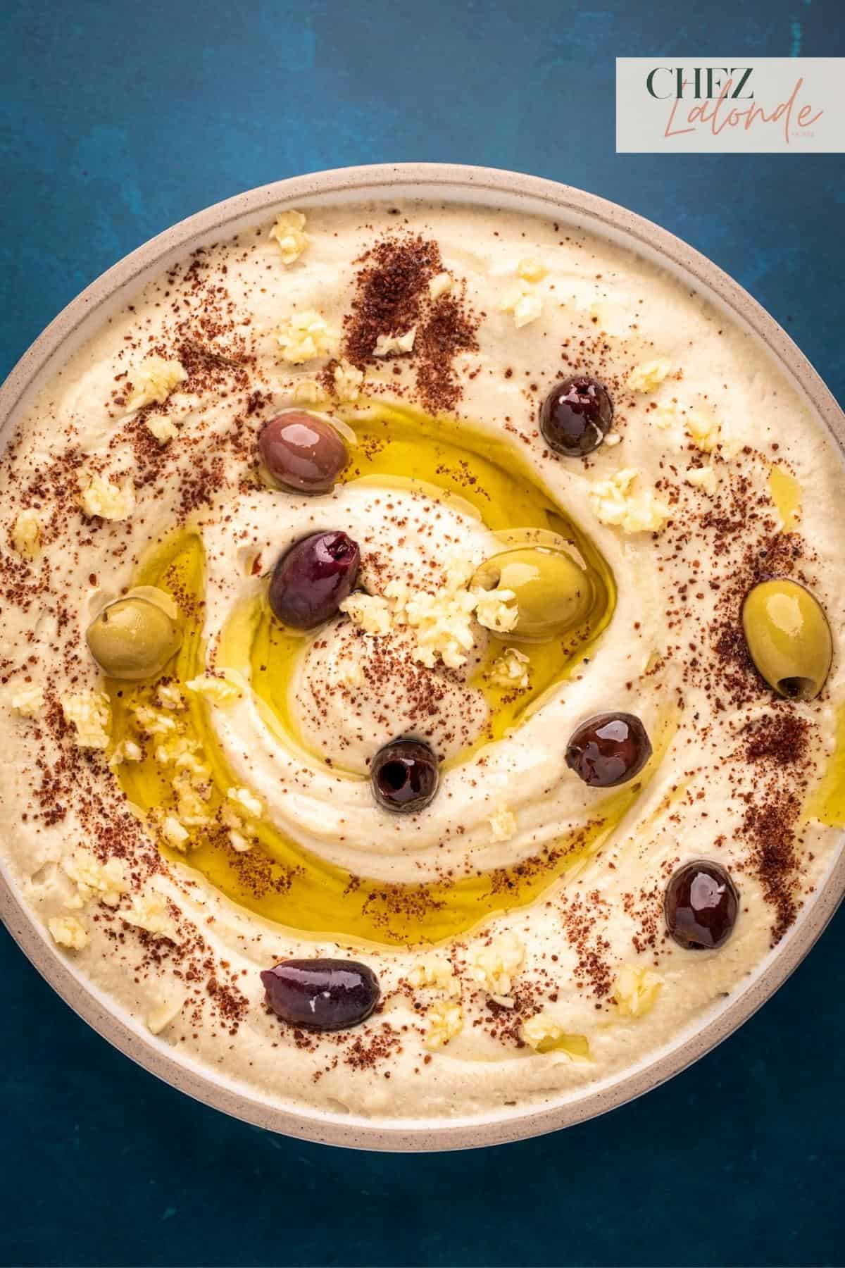 A bowl of hummus with olives and minced garlic. Sprinkled with Sumac and drizzled with extra virgin olive oil.