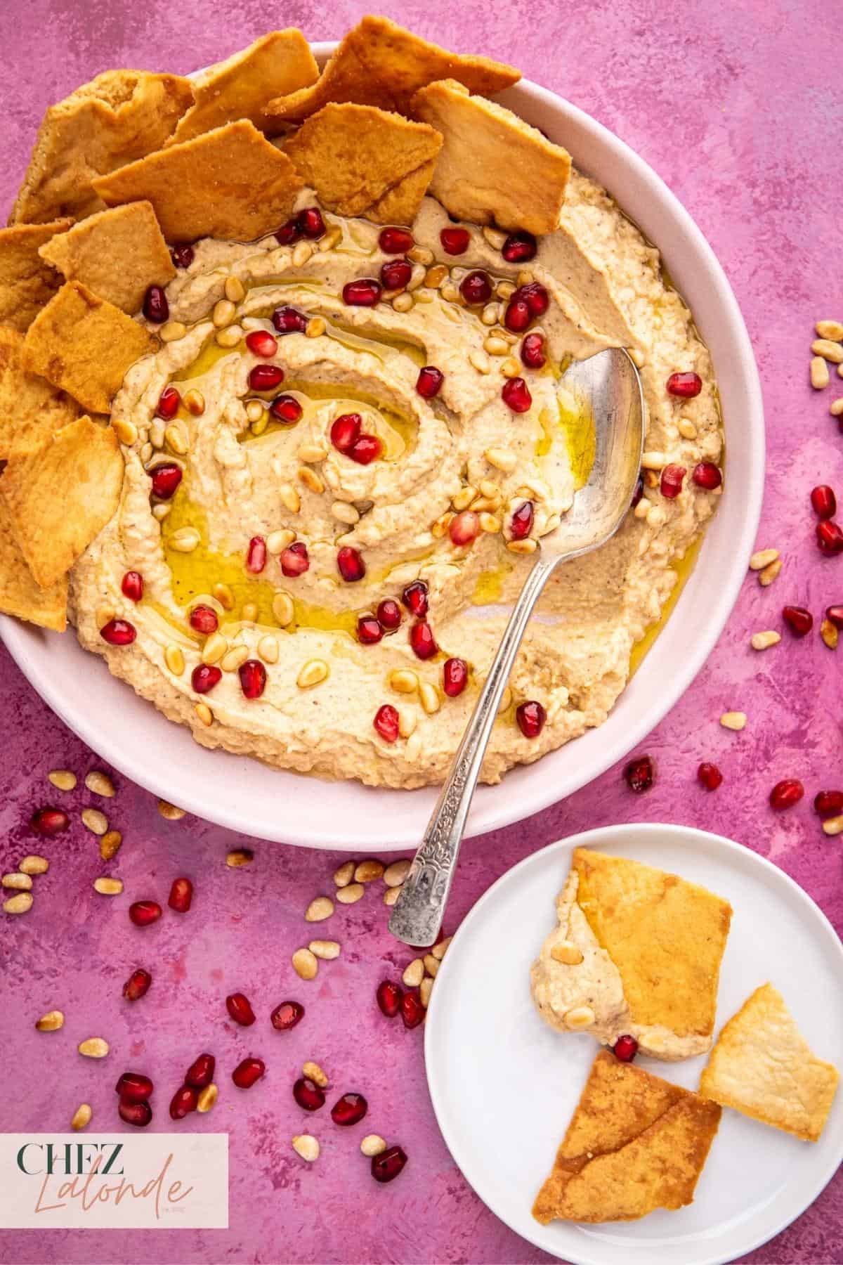 A bowl of baba ganoush serving with pita chips. Garnished with pomegranate seeds, olive oil, and roasted pine nuts. 