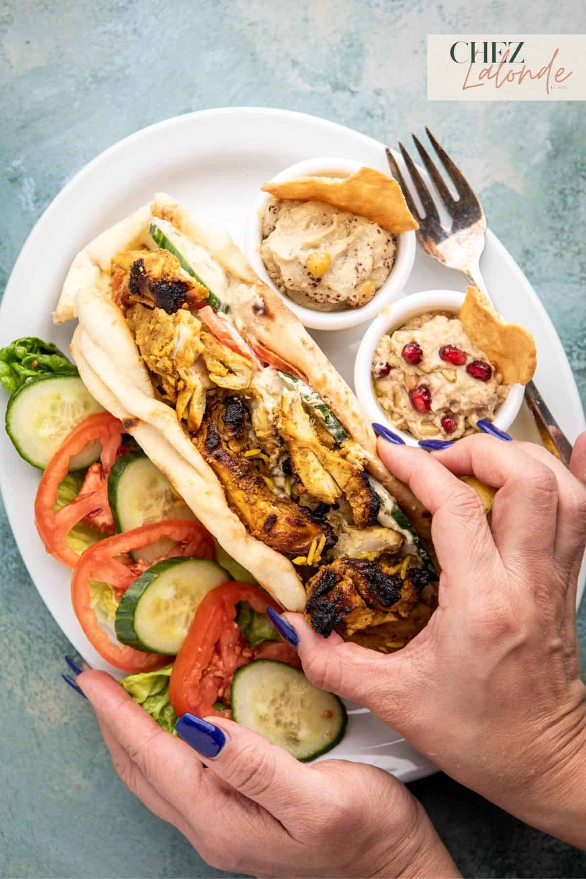 A hand is holding a chicken shawarma wrap. Serving with fresh veggies, hummus, and baba ganoush.