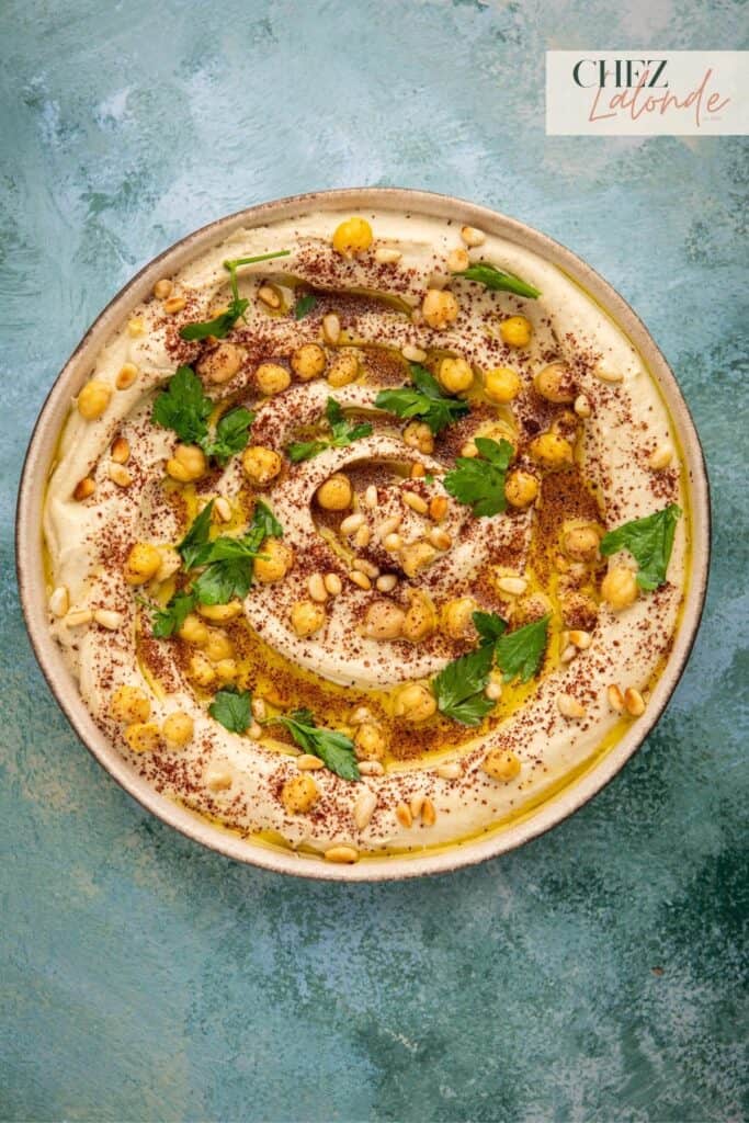 A bowl of hummus garnished with fresh chickpeas, toasted pine nuts and chopped parsley.