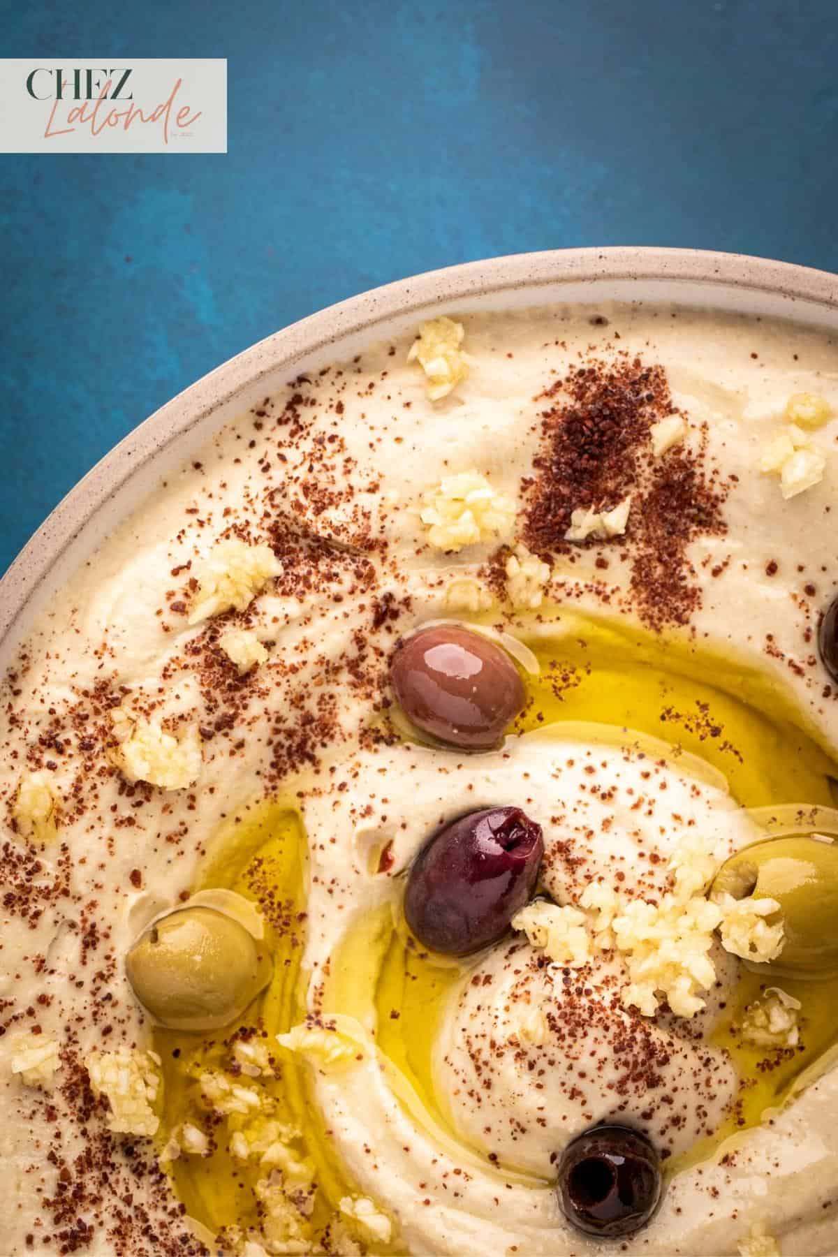 A bowl of hummus made with Vitamix.  Garnished with olives and minced garlic. Drizzled with olive oil. 