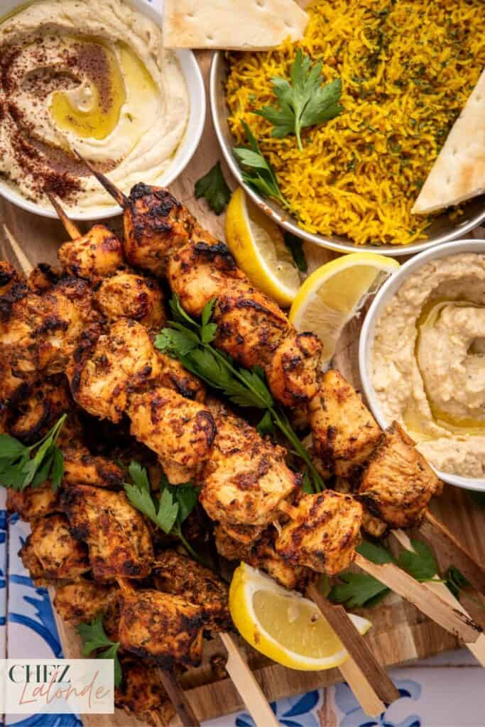 Shish tawook skewers on a wooden board pair with side dishes such as yellow rice, hummus, and baba ganoush.
