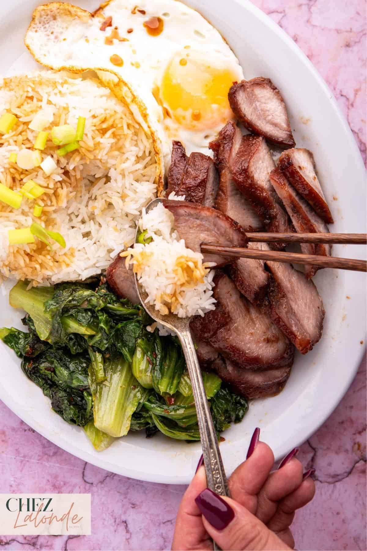 A woman is eating a Sous Vide Char Siu rice platter with Chinese BBQ pork, steamed rice and veggies and fried egg. 