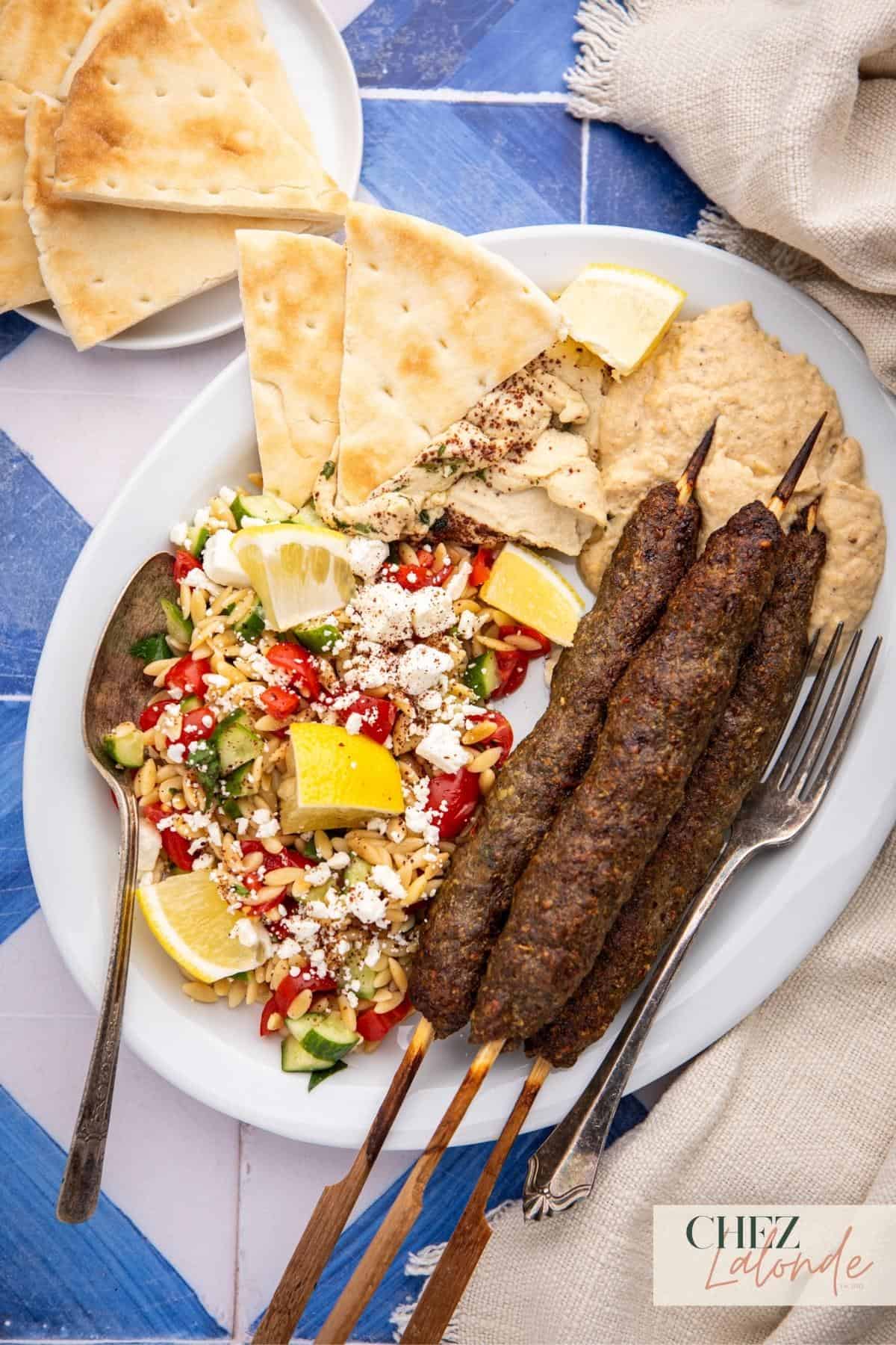 A plate contains 3 air fryer Kofta kebab skewers with Orzo salad and pita bread. Also there are side of Hummus and Baba ganoush for dipping the pitas. 