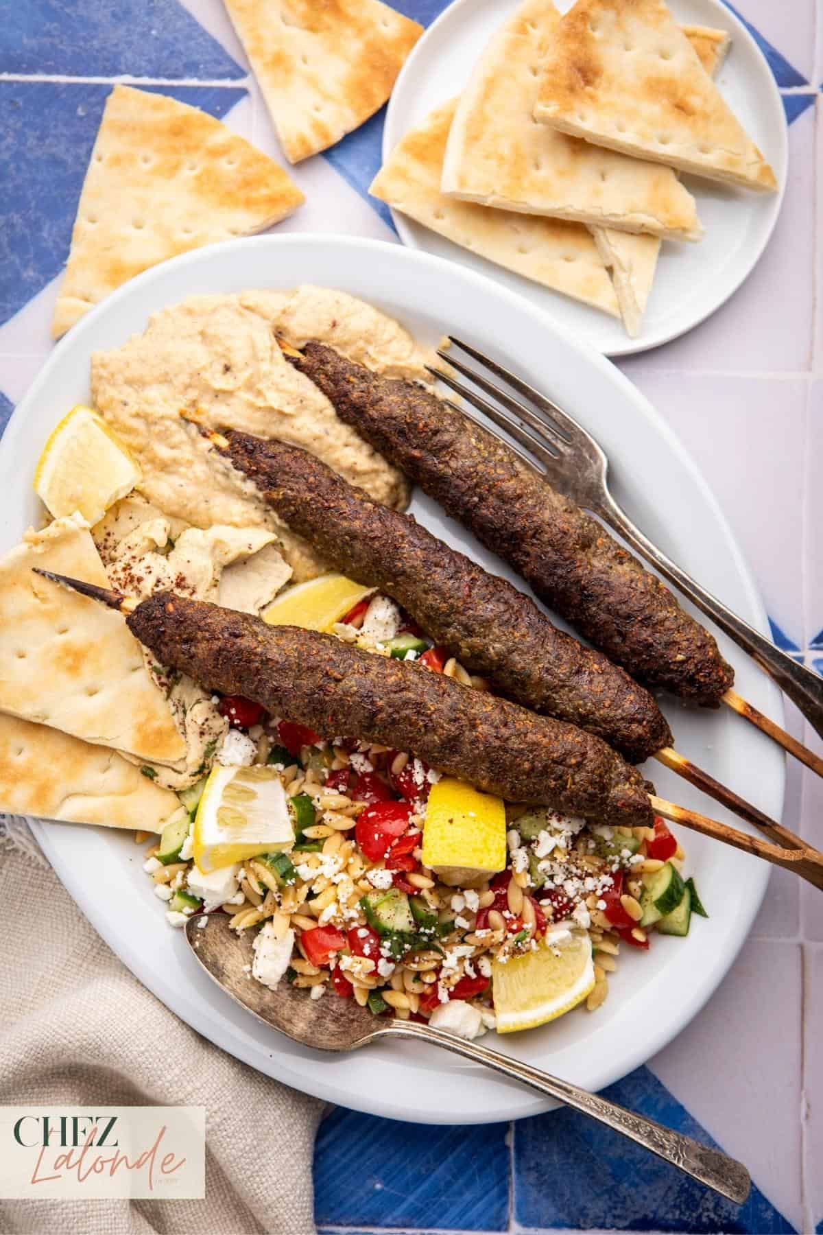 A plate with 3 skewers of Kofta kebabs served with Orzo salad, baba ganoush, hummus, and pita. 